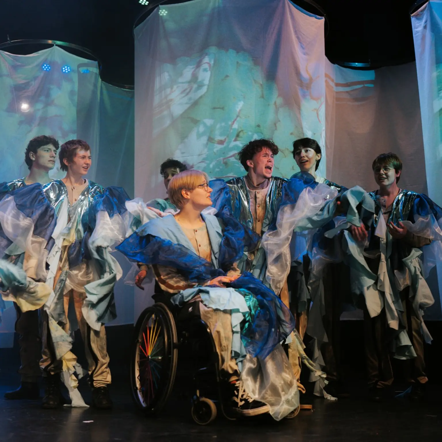 Een groep performers staat op een theaterpodium in fantasierijke kostuums van blauwe, witte en zilverkleurige stoffen. Centraal zit een persoon in een rolstoel, omringd door andere spelers die lijken te zingen of acteren. Op de achtergrond hangen transparante doeken waarop kleurrijke projecties worden getoond, terwijl blauw podiumlicht de scène een dromerige sfeer geeft.