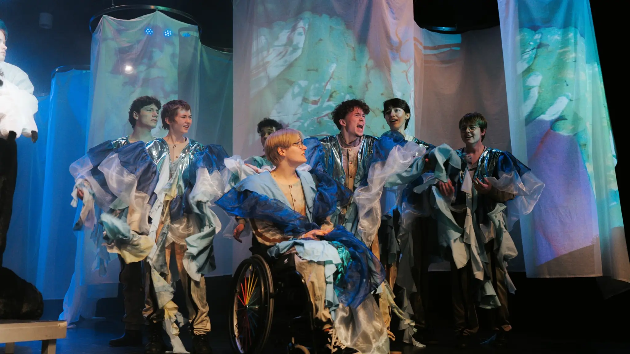 Een groep performers staat op een theaterpodium in fantasierijke kostuums van blauwe, witte en zilverkleurige stoffen. Centraal zit een persoon in een rolstoel, omringd door andere spelers die lijken te zingen of acteren. Op de achtergrond hangen transparante doeken waarop kleurrijke projecties worden getoond, terwijl blauw podiumlicht de scène een dromerige sfeer geeft.