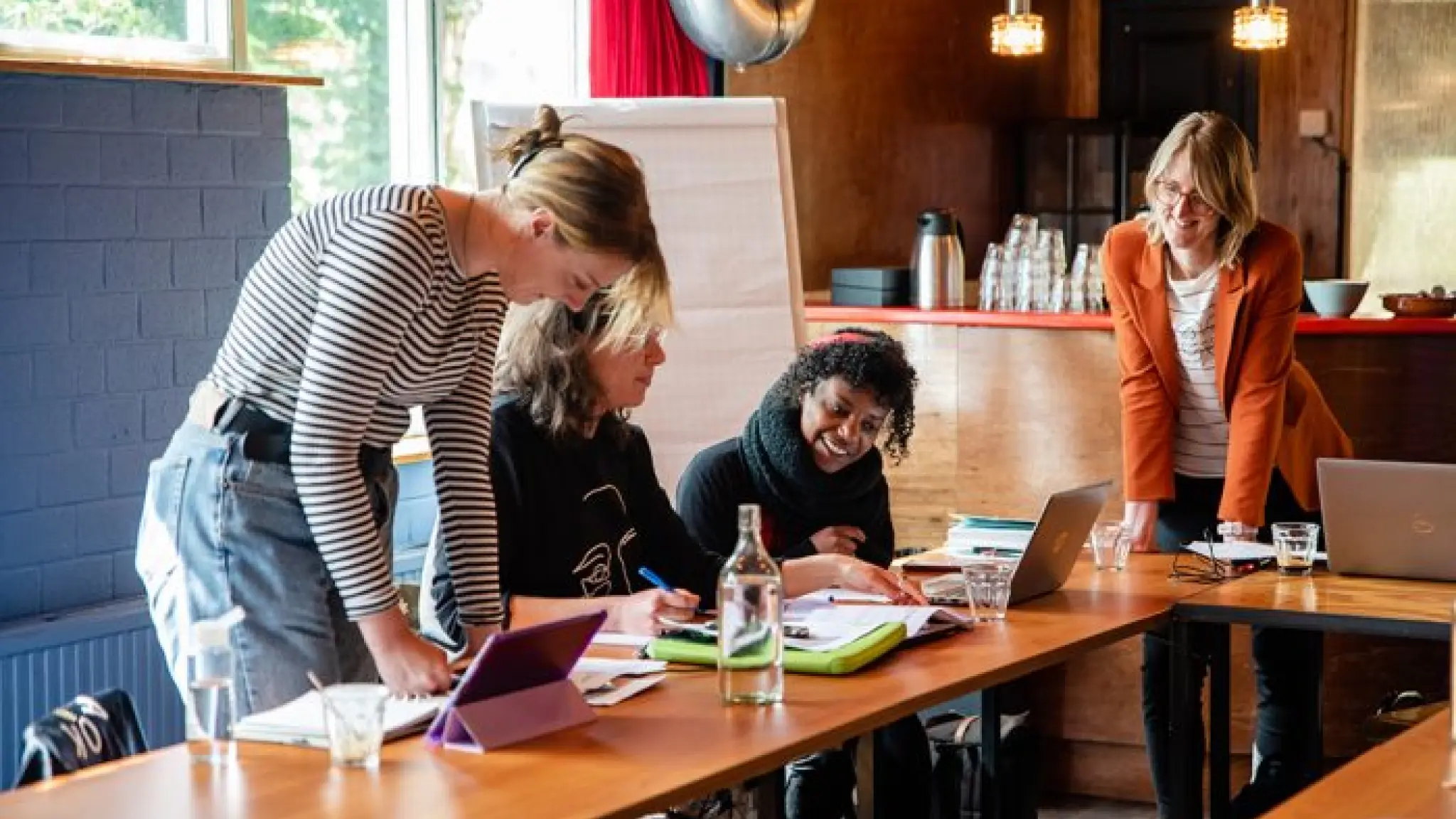 Vier mensen volgen een workshop. Ze zitten en staan aan een paar tafels en hebben laptops, schrijfblokken en glazen water voor zich.