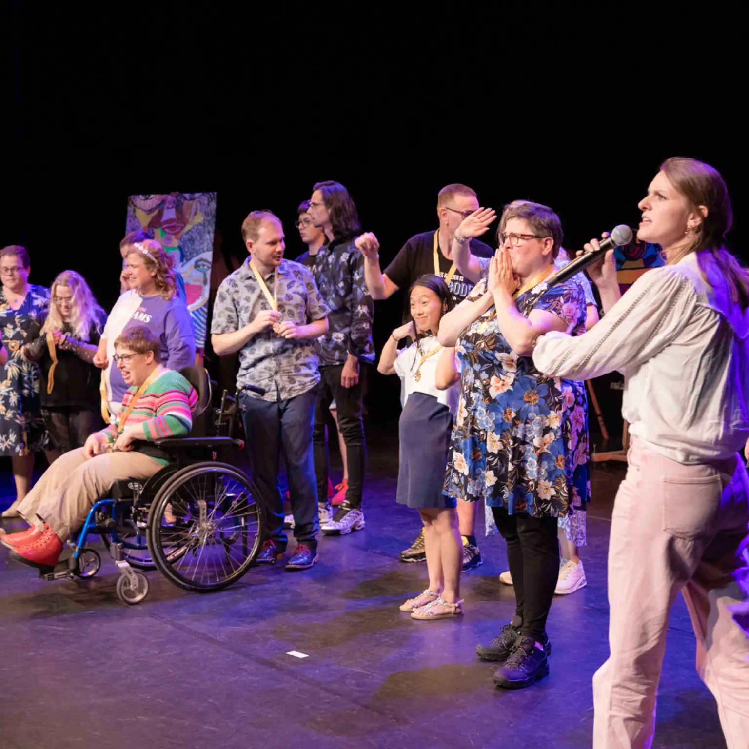 Een diverse groep deelnemers aan een theaterproject staat op het podium. Een presentator vraagt om applaus voor de groep. Een van de deelnemers zit in een rolstoel. Ze krijgen allemaal een medaille omgehangen.