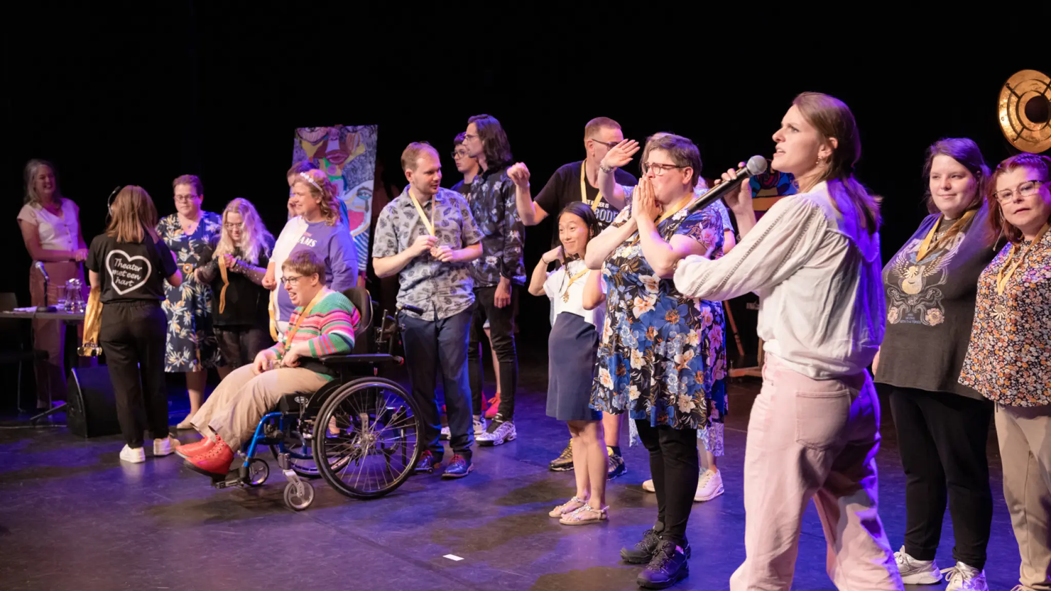 Een diverse groep deelnemers aan een theaterproject staat op het podium. Een presentator vraagt om applaus voor de groep. Een van de deelnemers zit in een rolstoel. Ze krijgen allemaal een medaille omgehangen.